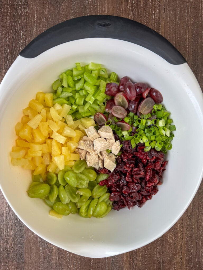 Mixing bowl with chopped celery, halved red and green grapes, sliced green onions, dried cranberries, pineapple tidbits and small cubes of chicken.