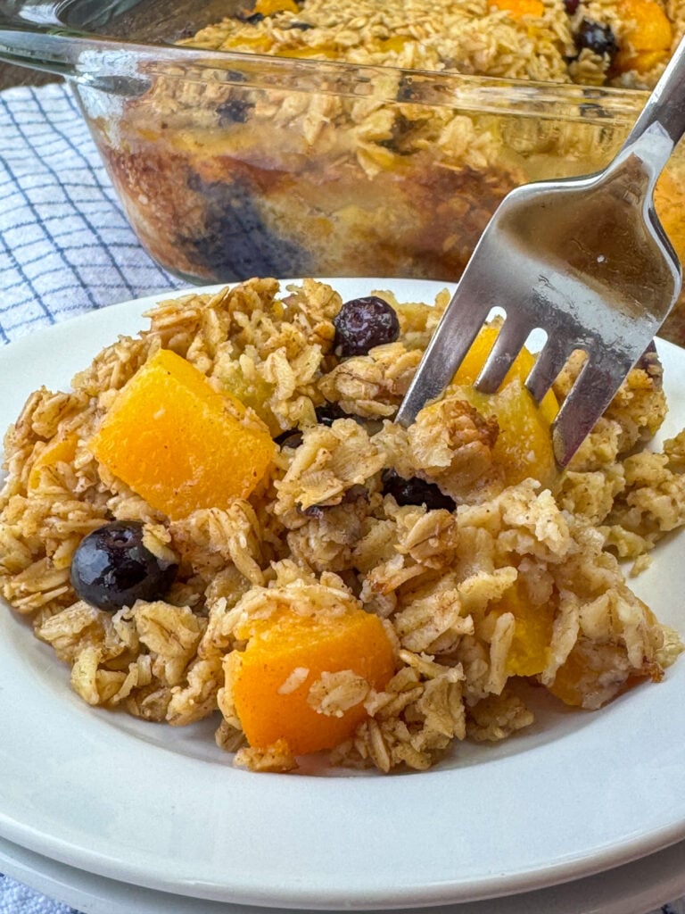 Close up delicious baked oatmeal on a fork with plenty of blueberries and chopped peaches on a plate with the full pan in the background.