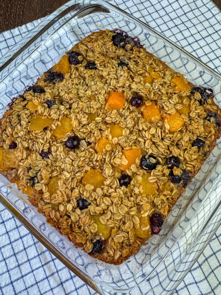 Overhead view of a square dish with golden brown baked oatmeal filled with blueberries and peaches on a blue and white napkin.