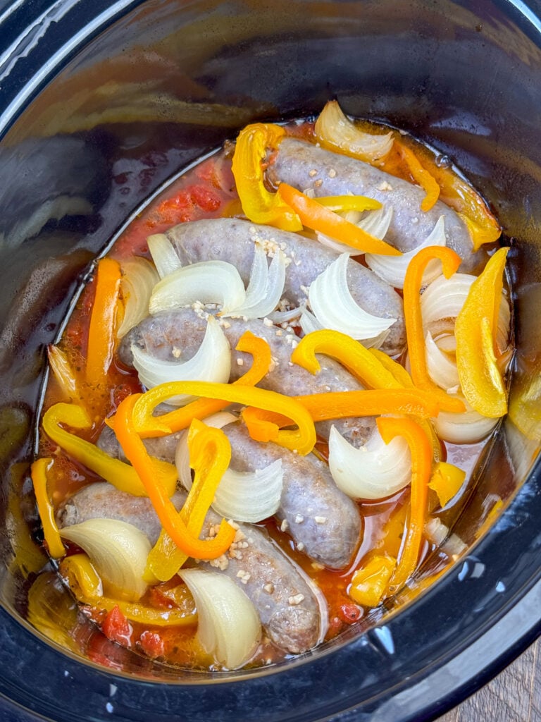 Brats cooking in beer with onions and pepper slices in a crock pot.