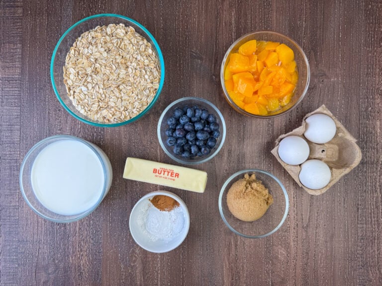 Ingredients to make baked oatmeal are old fashioned oats, peaches, blueberries, eggs, brown sugar, butter, milk, cinnamon, baking powder and salt on a wooden counter top.