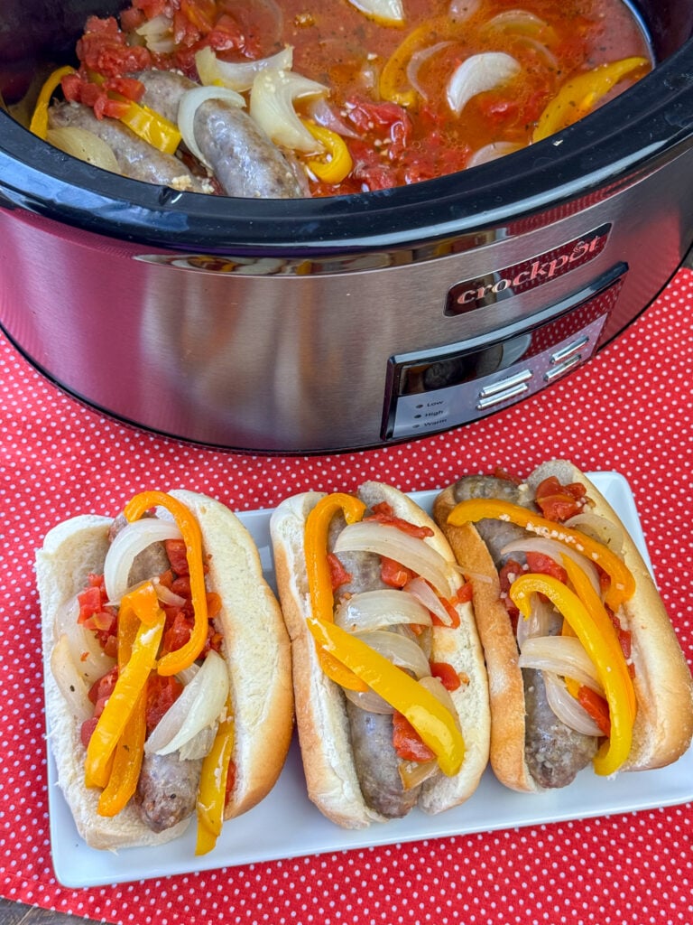 Overhead view of delicious brats in a crock pot and some on a tray in front in buns.