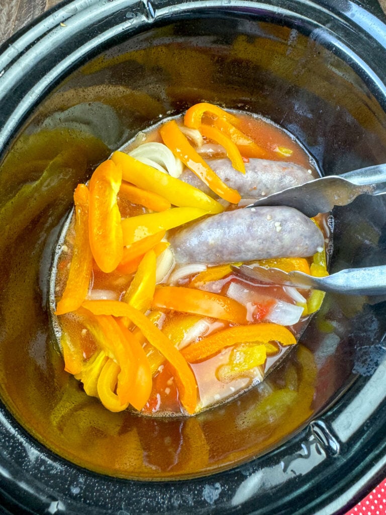 Tongs pulling cooked brats out of peppers and liquid in a black crock.