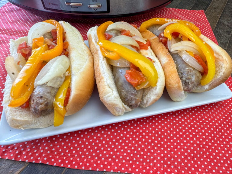 Side view of Brats in buns with peppers, onions and tomatoes on top and the buns are on a white tray.