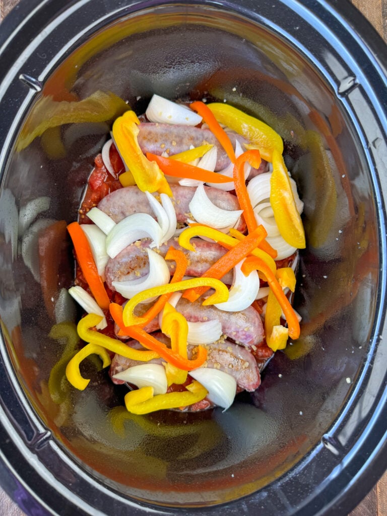 Sliced bell pepper and onion spread over minced garlic on brats in a large black slow cooker.