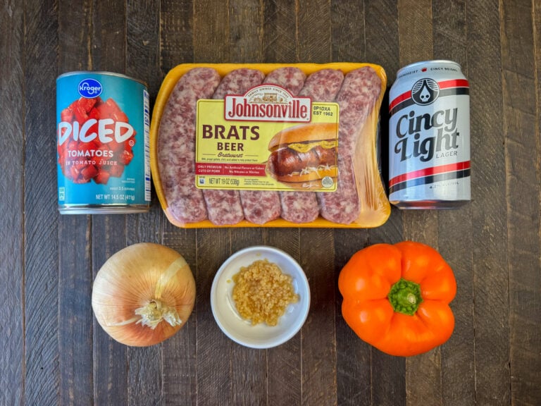 Ingredients to make brats in a slow cooker are diced tomatoes, brats, can of beer, bell pepper, minced garlic and an onion on a wooden counter top.