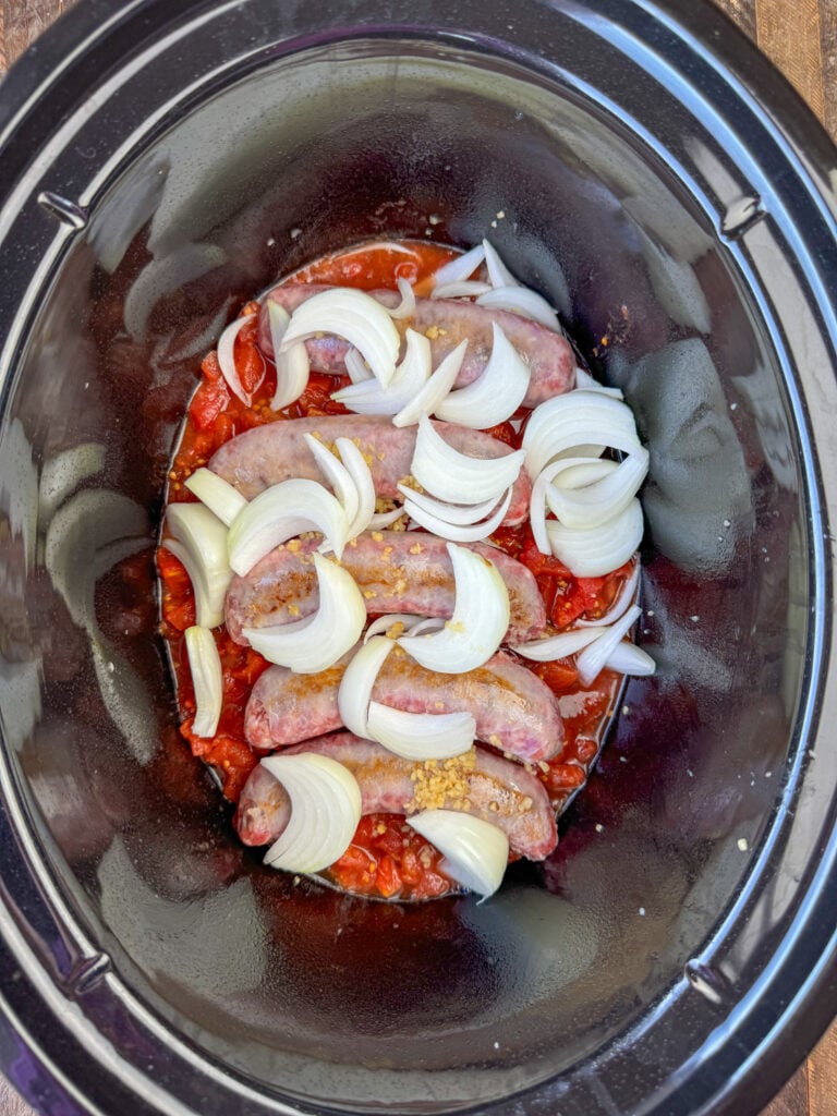 Brats on a bed of diced tomatoes and beer with onions and garlic on top in a large black slow cooker.