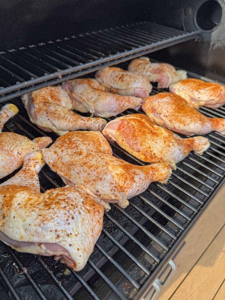 Raw chicken quarters with seasoning rubbed in arranged fat side up on a smoker.