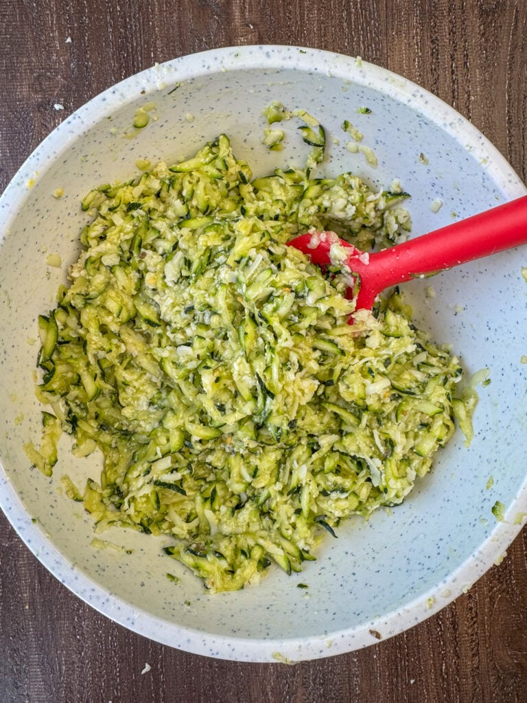 Spatula stirring grated zucchini with cheese and eggs in a mixing bowl.