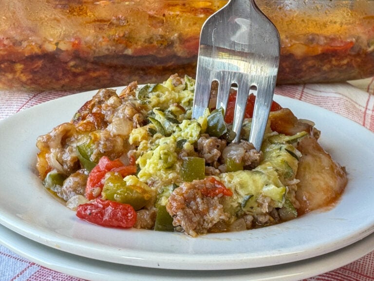 Flavorful sausage and zucchini casserole baked with a few onions, tomatoes and green peppers and coated with melted cheese on a plate with a fork ready to take a bite.