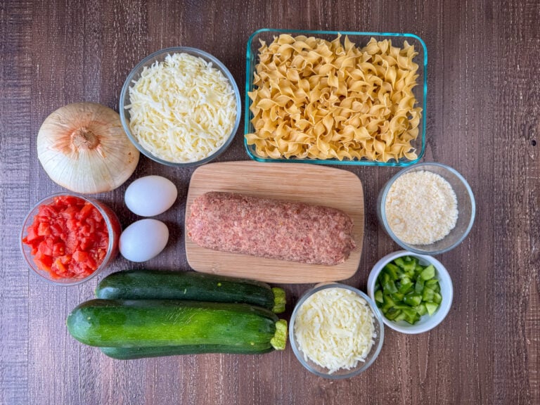 Ingredients in zucchini and sausage casserole are egg noodles, parmesan cheese, Italian sausage, diced green pepper, Italian cheese, zucchini, eggs, tomatoes, onion and mozzarella cheese on a wooden counter top.