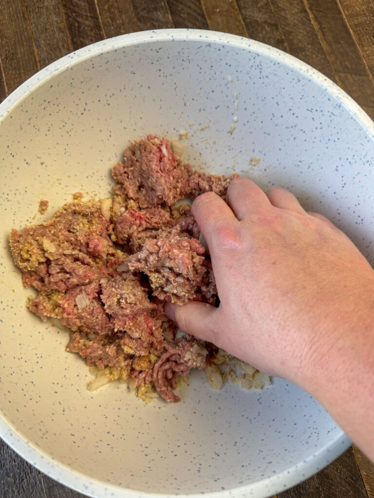 Hand mixing breadcrumbs, seasonings and onions into ground beef in a white speckled bowl.