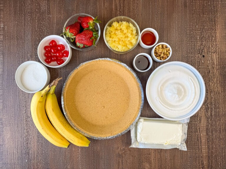 Ingredients to make Banana Split Pie are ripe bananas, prepared graham cracker crust, Cool Whip, cream cheese, chocolate syrup, chopped nuts, vanilla, crushed pineapple, strawberries, cherries and sugar on a wooden counter top.