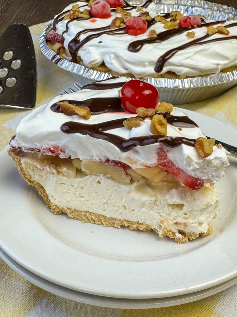 Side view of a slice of no bake banana split pie with the full dessert in the background showing a thick cheesecake like base, layers of fruit and simple garnishes on top.