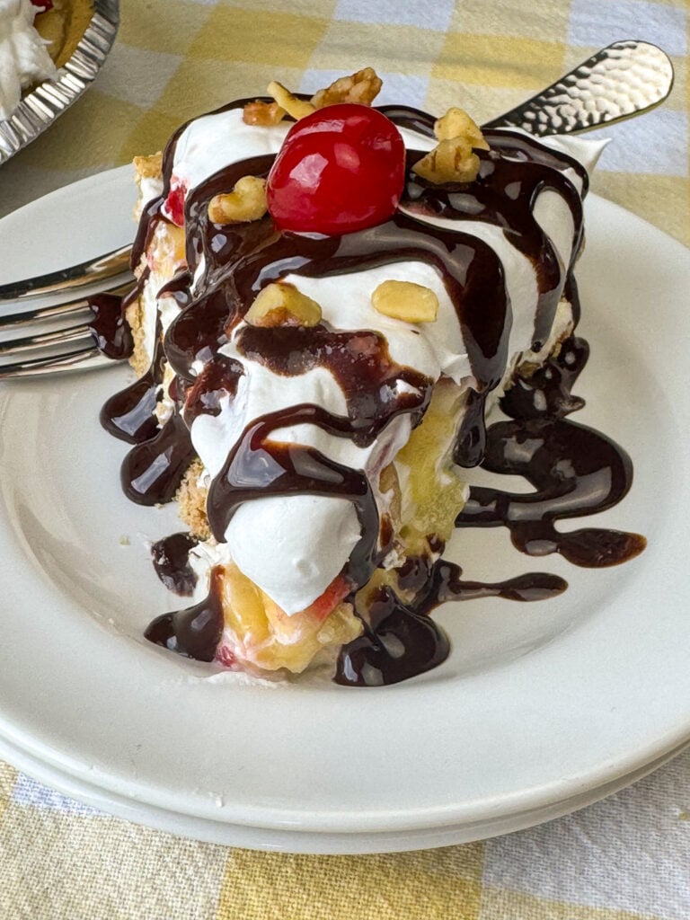 Chocolate syrup drizzled generously over a slice of banana pie with cool whip, cream cheese, crushed pineapple and strawberries on a white plate with a fork in the background.