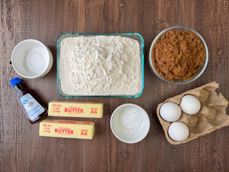 Ingredients needed to make caramel cookies are flour, dark brown sugar, eggs, baking soda, butter, vanilla and salt on a wooden counter top.
