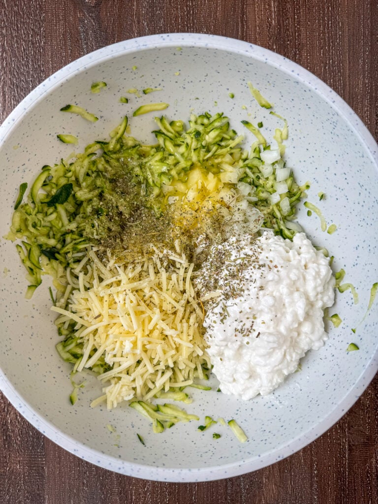 Mixing bowl with a generous amount of shredded zucchini, cottage cheese, chopped onion, Monterey Jack cheese and Italian seasoning sprinkled over the top.