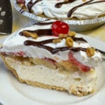 Close up of slightly frozen slice of banana split pie with a thick creamy layer topped with fruit and whipped topping then drizzled with chocolate and a cherry placed on top.