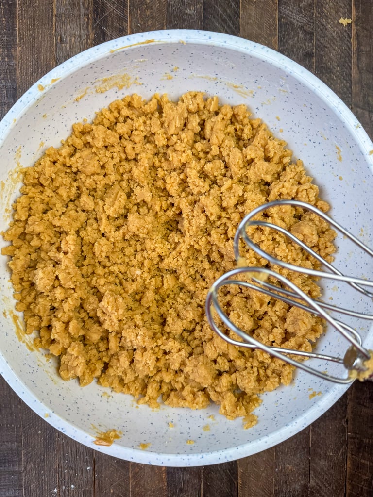 Mixer beaters resting above a mixing bowl with a rich, crumbly mixture of peanut butter, shortening, sugar, eggs and vanilla.