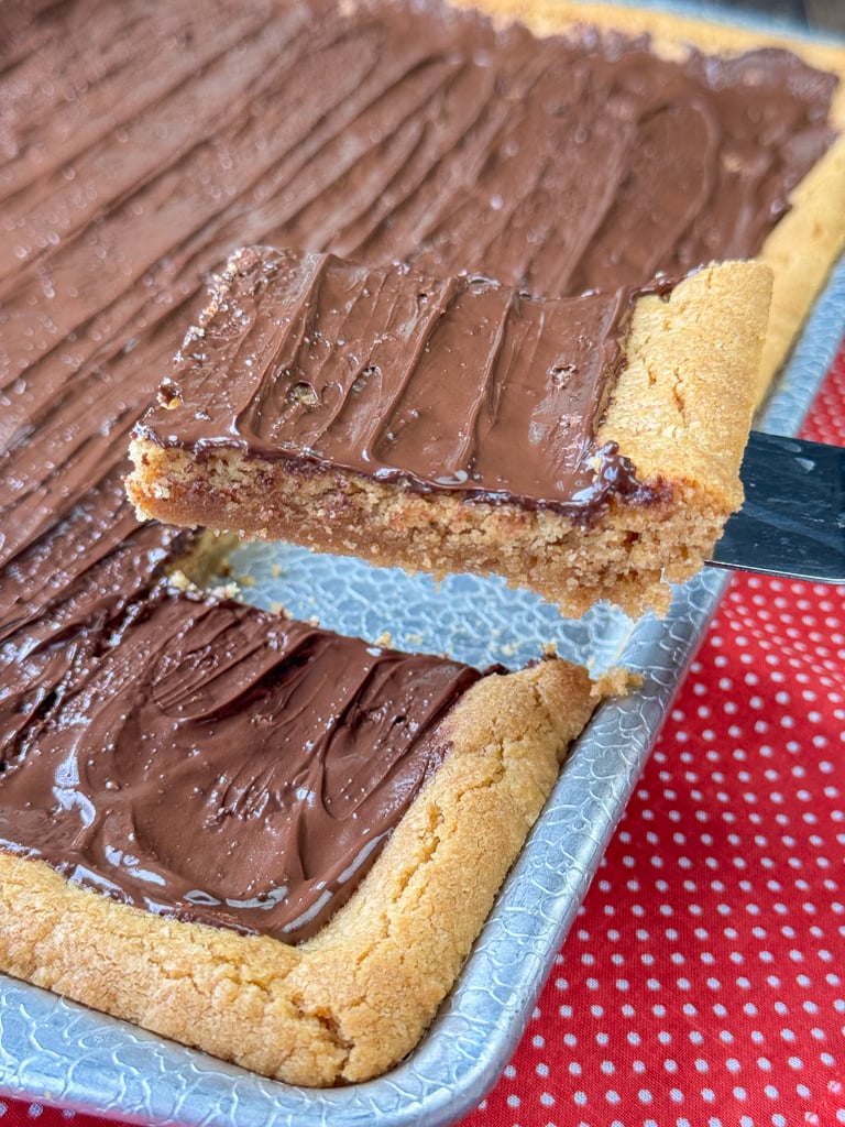 Scoop lifting a peanut butter bar with a thin layer of melted chocolate above the sheet pan full revealing a slightly gooey texture.