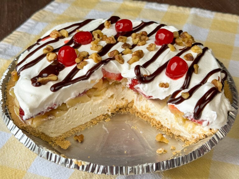 Foil pie pan with a chilled, creamy and delicious banana split pie layered on a graham cracker crust and topped with chocolate syrup, chopped nuts and cherries on a yellow and white napkin.