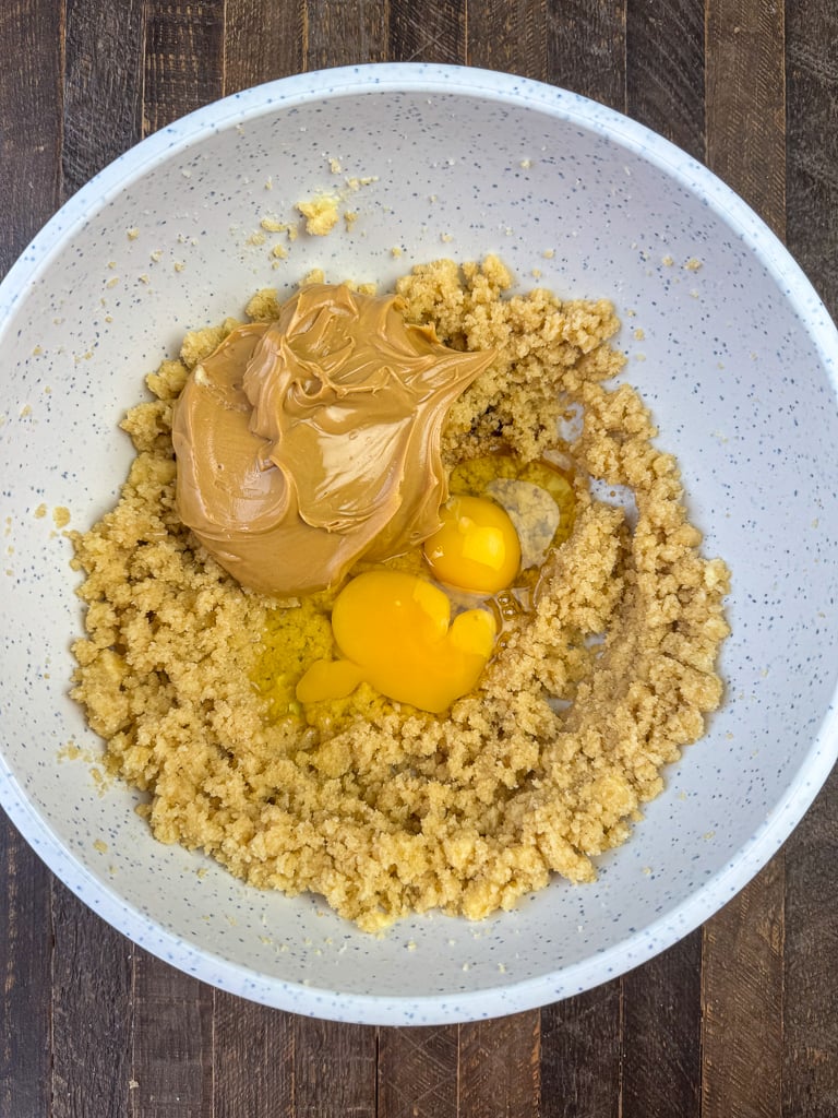 Peanut butter and eggs added to a bowl of peanut butter, flour and sugar crumbles.