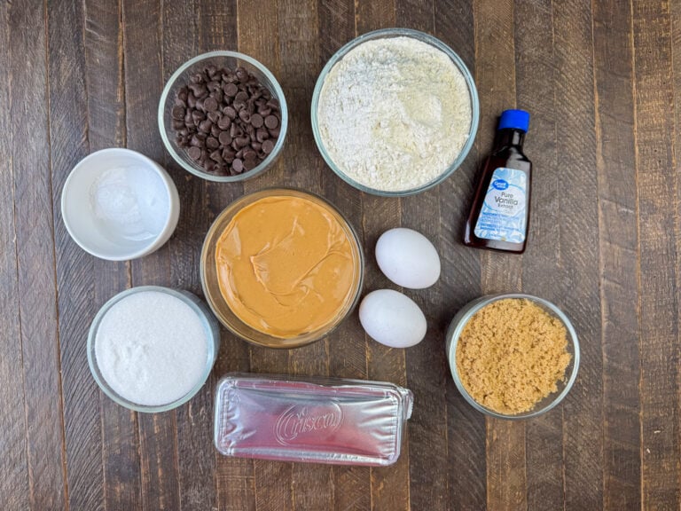 Ingredients to make peanut butter bars are flour, vanilla, brown sugar, eggs, peanut butter, shortening, sugar, baking soda and chocolate chips on a wooden counter top.