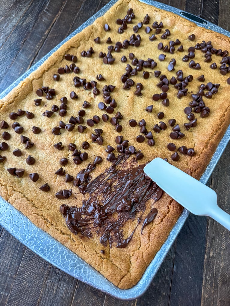 Spatula spreading chocolate chips over a sheet pan of warm peanut butter bars.