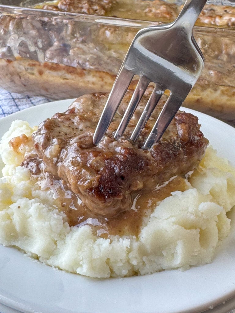 Poor man's steak made of hamburger resting on a generous bed of mashed potatoes with the fork ready to take a bite and the full dish of steaks in the background.