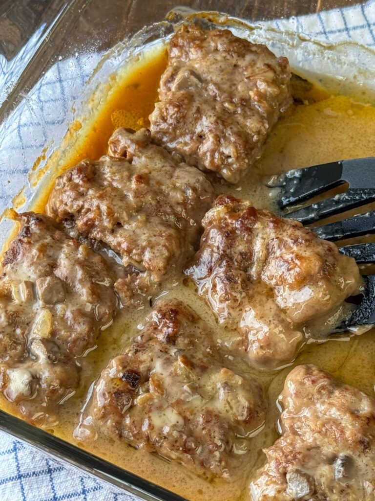 Spatula lifting an Amish hamburger steak out of a gravy filled baking dish full of steaks.