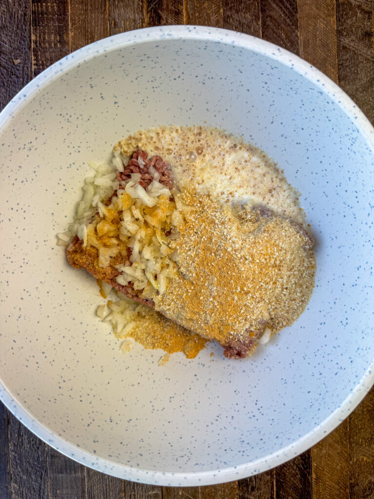 Seasoned salt, bread crumbs, chopped onion and milk poured over ground beef in a mixing bowl.