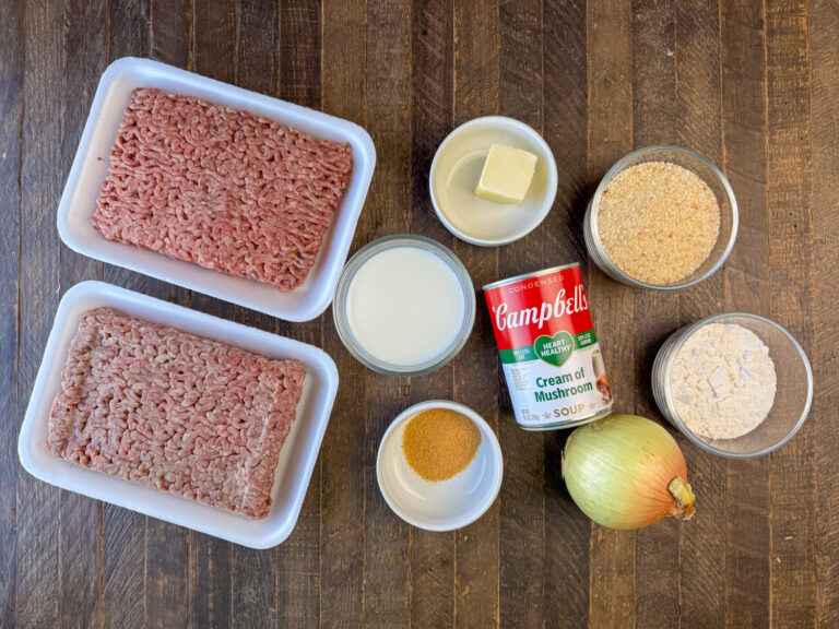 Ingredients to make poor mans steak are ground beef, milk, butter, seasoned salt, cream of mushroom soup, bread crumbs, flour and an onion on a wooden counter top.