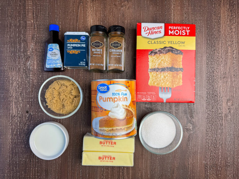 Ingredients to make Pumpkin Dump Cake are a yellow cake mix, can of pure pumpkin, sugar, butter, brown sugar, milk, cinnamon, nutmeg, pumpkin pie spice and vanilla on a wooden counter top.