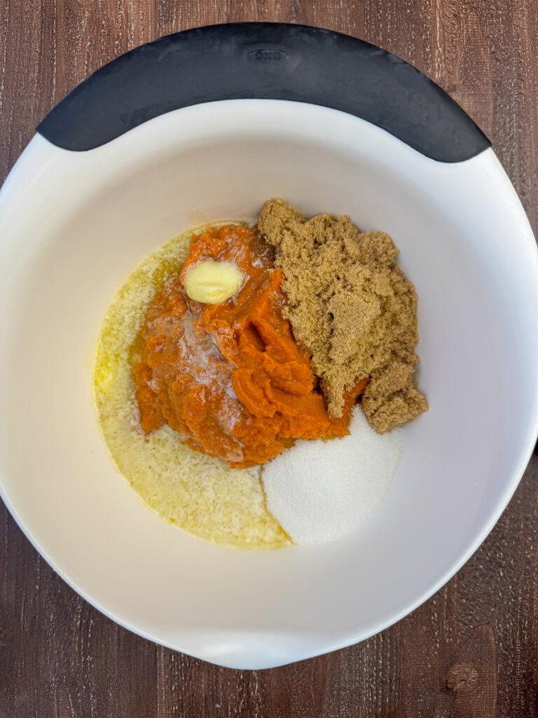 Brown sugar, granulated sugar and melted butter poured over a large can of pumpkin puree in a wooden mixing bowl.
