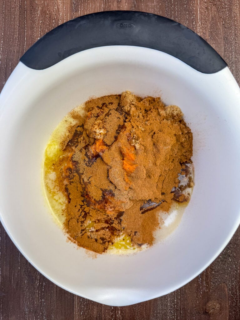 Pumpkin pie spice, nutmeg and cinnamon sprinkled over top of pure pumpkin, a yellow cake mix, with brown sugar, sugar, milk and melted butter on a wooden counter top.