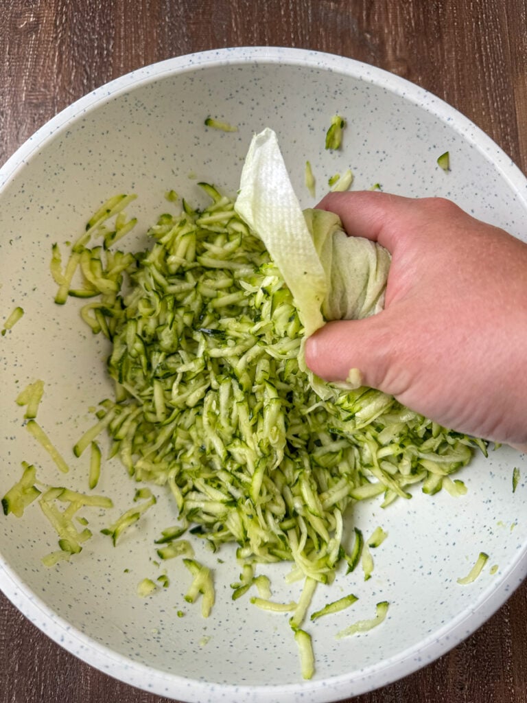 Hand squeezing shredded zucchini over a mixing bowl.