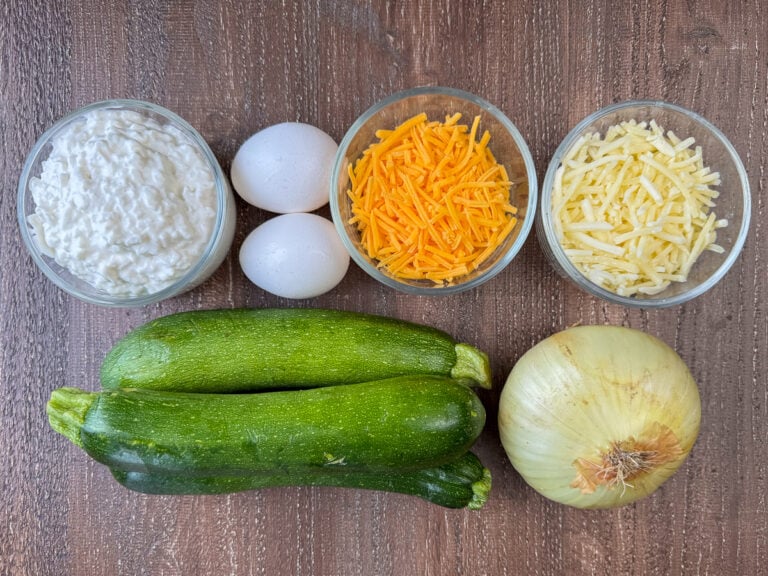 Ingredients to make a zucchini casserole are cottage cheese, eggs, cheddar cheese, white cheese, zucchini and onion on a wooden counter top.