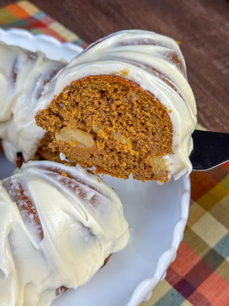 Spatula lifting a piece of apple pumpkin cake with cream cheese frosting up above the rest of the cake to serve.
