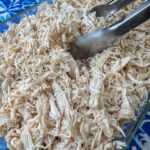 Tongs in the center of a dish full of baked and shredded chicken breasts.