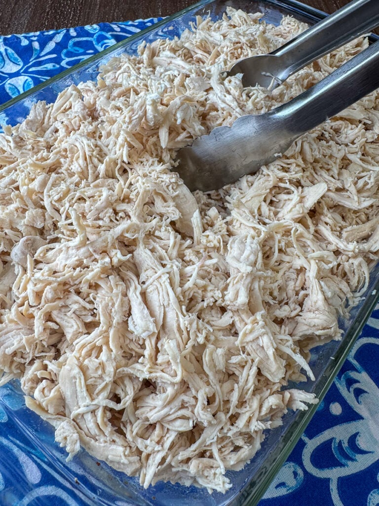 All white meat shredded chicken ready to serve with tongs in a glass casserole dish on a blue napkin.