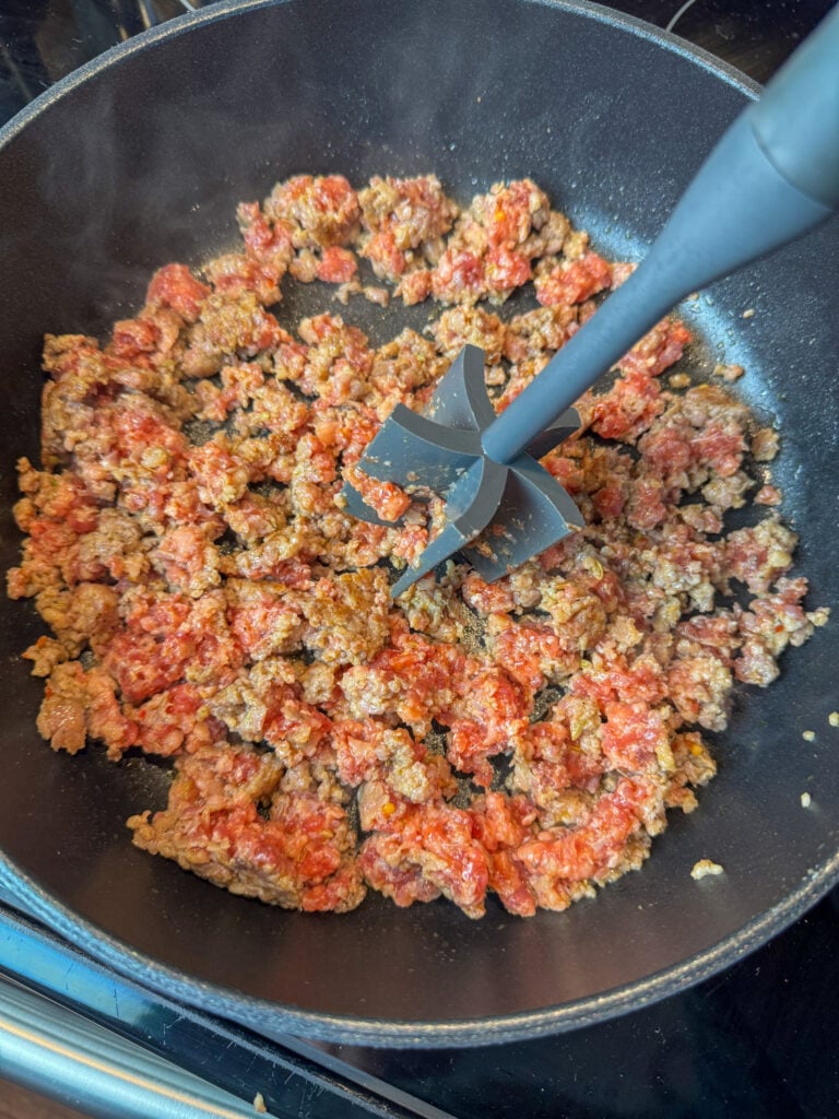Meat masher stirring breakfast sausage as it cooks in a deep skillet.