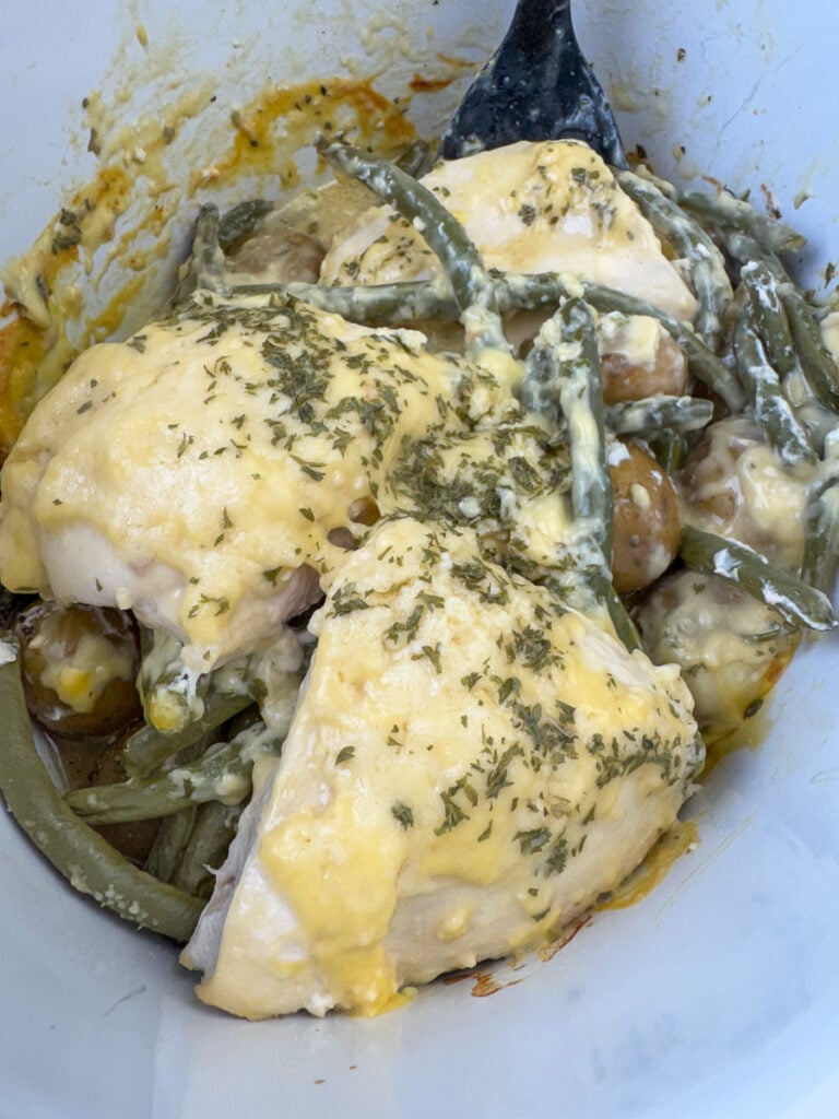Serving spoon scooping up baby gold potatoes, green beans and tender pieces of chicken breast up out of a white slow cooker.