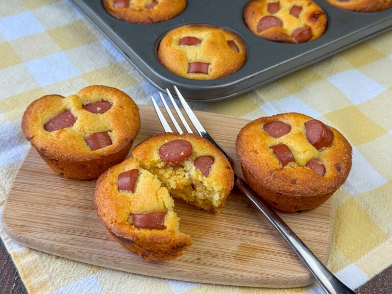 Fork in the center of a little wooden board with corn muffin hot dogs and one is cut in half ready to eat.