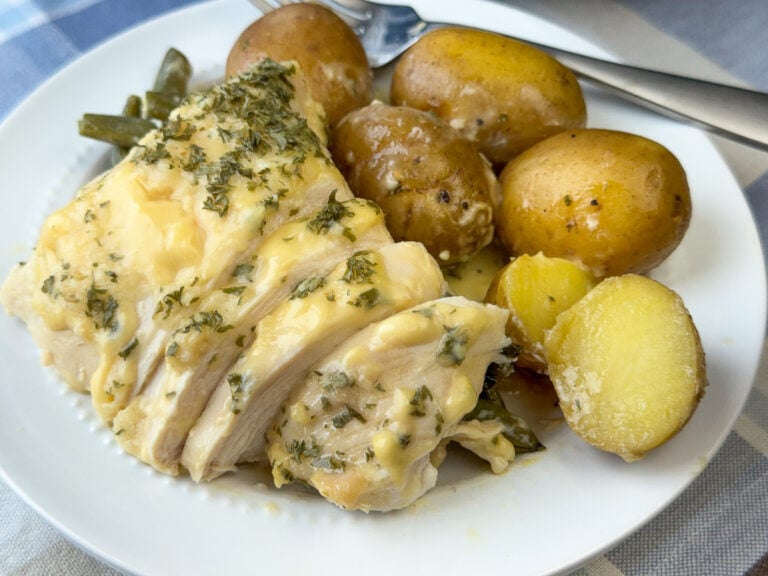 Tender sliced chicken breast laid on tender green beans and baby potatoes on a white plate with a fork in the background.