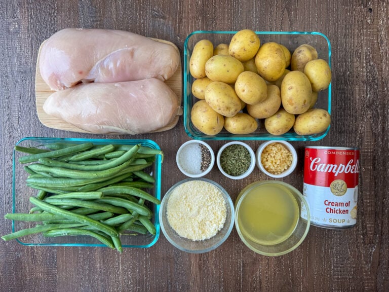 Ingredients to make crock pot chicken and potatoes are chicken breasts, baby potatoes, can of cream of chicken soup, chicken broth, minced garlic, parsley, parmesan cheese, salt, pepper and green beans on a wooden counter top.