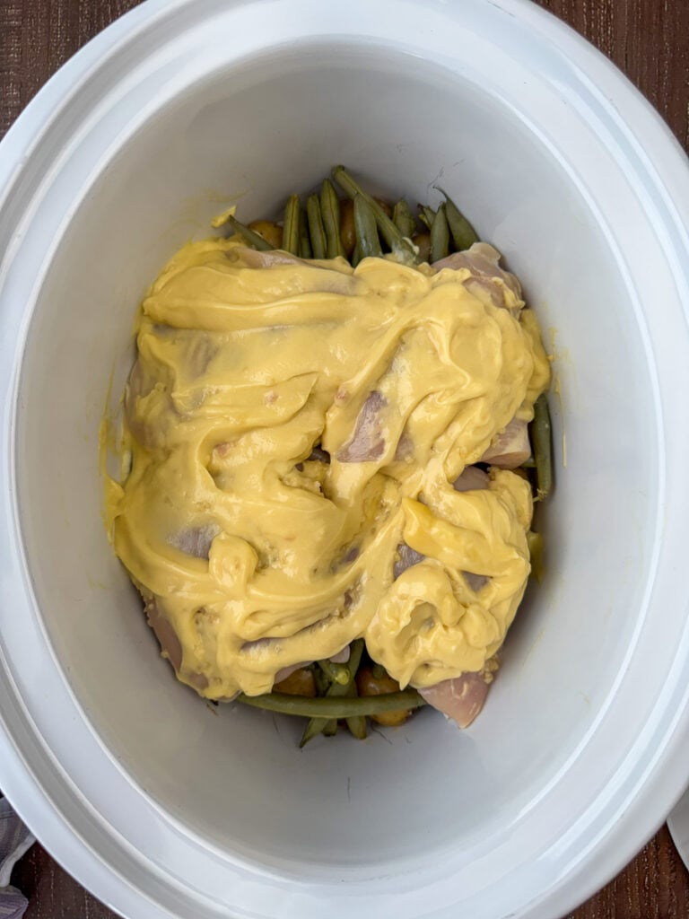 Thick layer of cream of chicken soup spread over chicken, green beans and potatoes in a slow cooker.