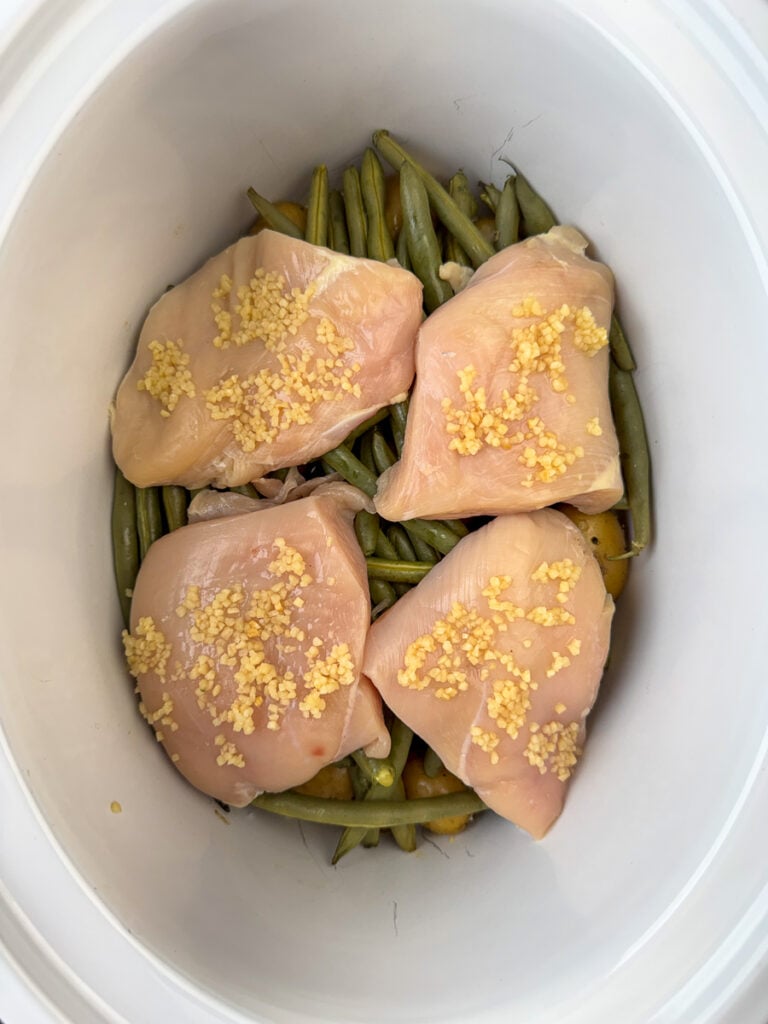Generous amount of minced garlic spread over chicken breasts that are resting on potatoes and green beans in a crock pot.