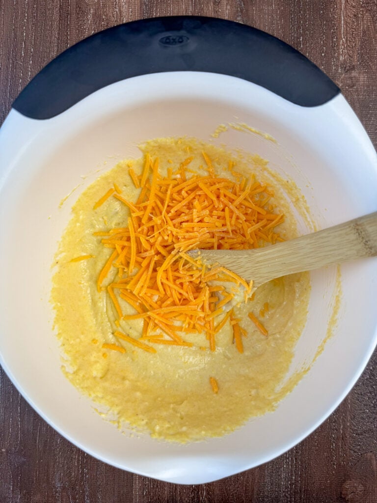 Shredded cheddar cheese added to corn muffin mix in a white bowl.