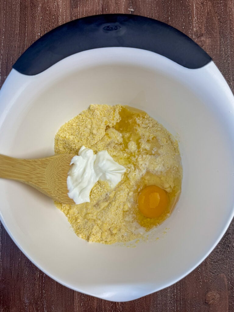 Wooden spoon stirring sour cream into a corn muffin mix along with oil and an egg in a white bowl.