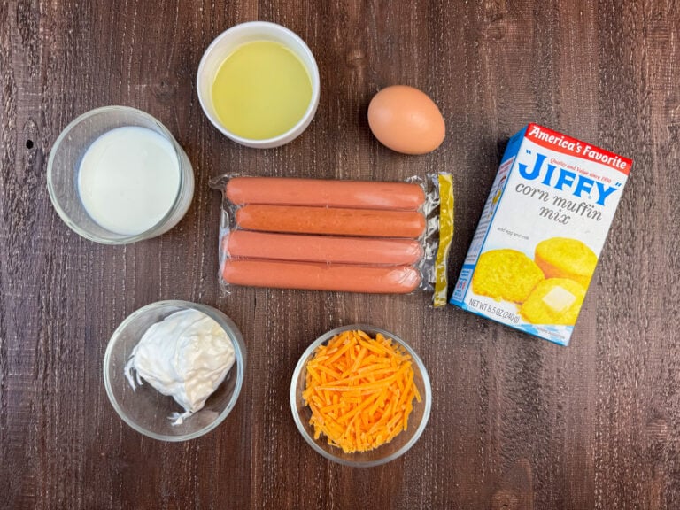 Ingredients to make hot dog corn muffins are milk, vegetable oil, hot dogs, an egg, Jiffy corn muffin mix, cheddar cheese and sour cream on a wooden counter top.